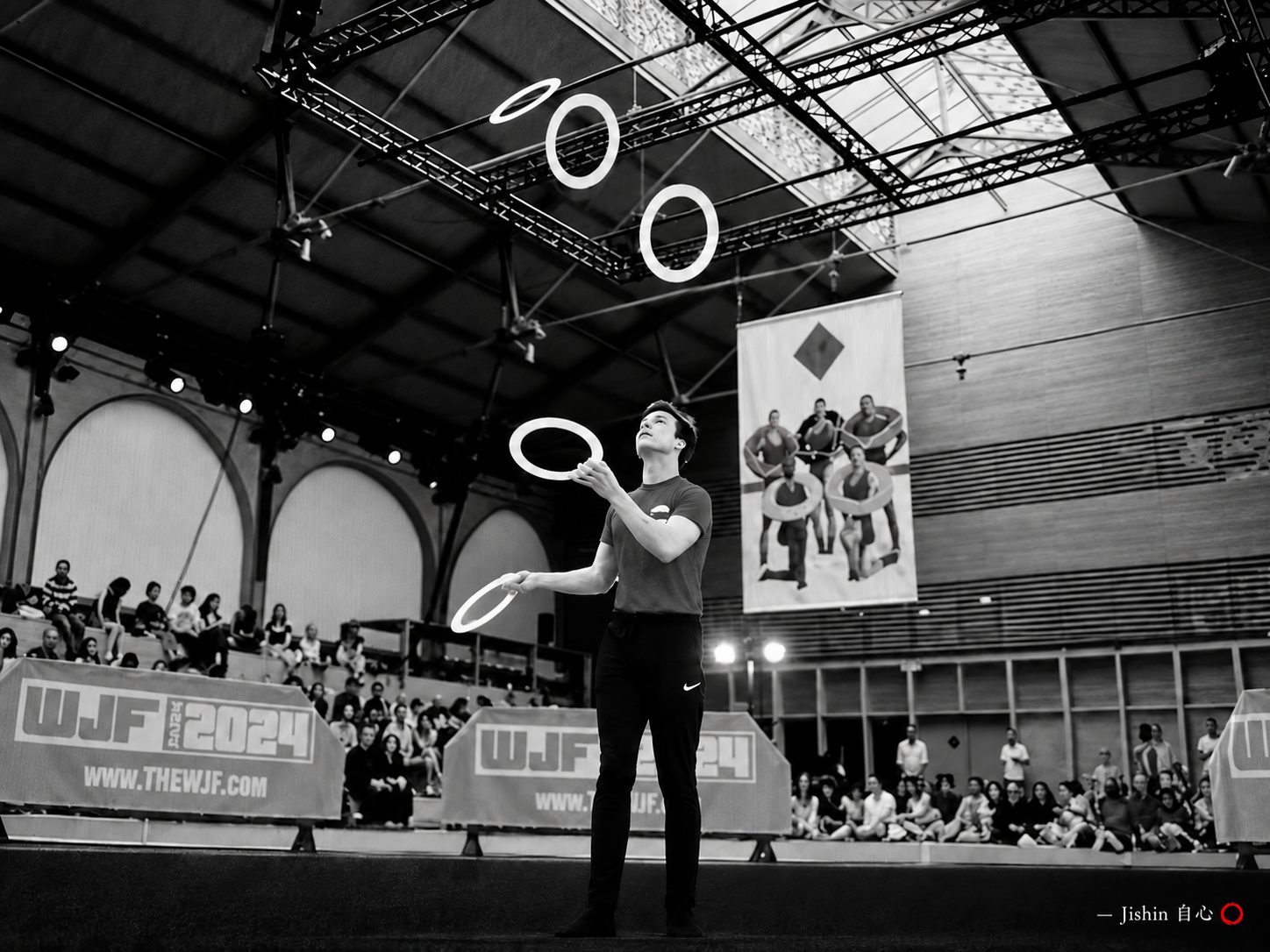 A juggler balances bright rings in the air before a packed audience, in black and white with one red ensō